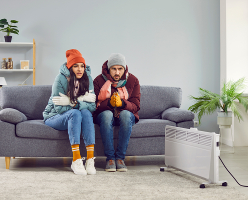 Freezing people in winter jackets sitting on sofa at home. Sad warmly closed husband abd wife feeling cold and trying to warm up. Heating problems, power crisis concept