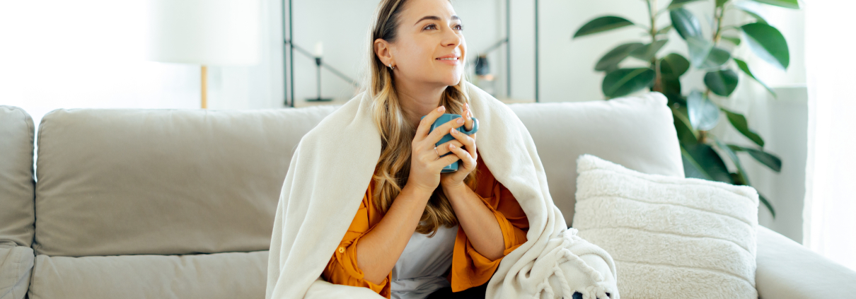 Beautiful caucasian young woman in yellow shirt sits on a sofa at living room wrapped in a cozy blanket, holding a blue mug tea or coffee, she looks relaxed and happy, spending weekend at home, smile