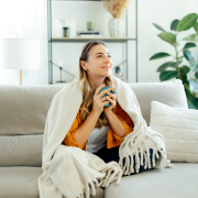 Beautiful caucasian young woman in yellow shirt sits on a sofa at living room wrapped in a cozy blanket, holding a blue mug tea or coffee, she looks relaxed and happy, spending weekend at home, smile