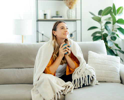 Beautiful caucasian young woman in yellow shirt sits on a sofa at living room wrapped in a cozy blanket, holding a blue mug tea or coffee, she looks relaxed and happy, spending weekend at home, smile